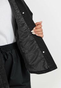 Mustat tilkkutakit, joissa on korhuva ulkopinta, napit ja sileä vuori. Näkyvä käsi pitää auki hihasta.