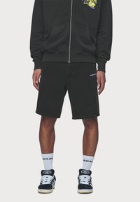 Mustat puuvillahousut suoralinjaisella leikkauksella, joissa on valkoinen logo vasemmassa sääressä. Yhdistetty mustan vetoketjuhupparin ja mustien lenkkareiden kanssa.