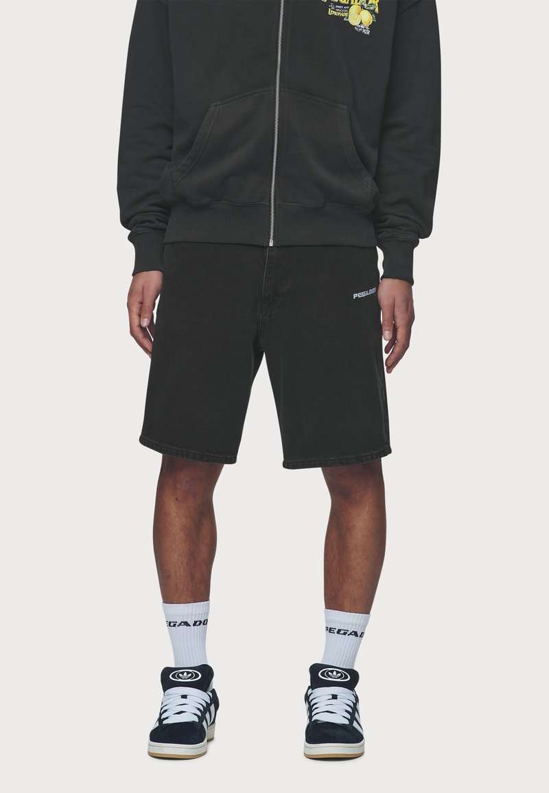 Mustat puuvillahousut suoralinjaisella leikkauksella, joissa on valkoinen logo vasemmassa sääressä. Yhdistetty mustan vetoketjuhupparin ja mustien lenkkareiden kanssa.