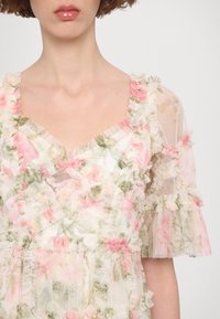 Femme portant une robe en tissu transparent à volants, à motif floral avec des accents roses et verts, manches courtes bouffantes, vue du cou à la taille.