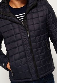 Laivastonsininen tikattu toppatakki, jossa on kaksi vetoketjullista sivutaskua ja korkea kaulus. Takissa on kiiltävä, sileä pinta ja kontrastiväriset mustat vetoketjut.