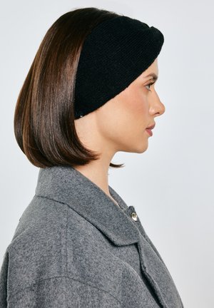 Moteris su tiesiais rudais plaukais, vilkinti juodą megztą galvos juostą ir pilką vilnonį paltą, profilio vaizde priešais paprastą foną.