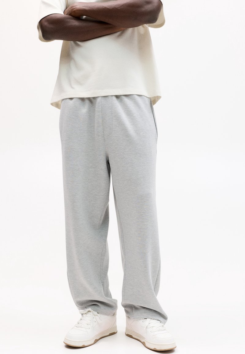 Personne portant un pantalon de survêtement ample gris clair, des baskets blanches et un t-shirt crème à manches courtes, debout, les bras croisés.