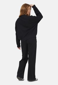 Femme aux longs cheveux roux se tient de dos, portant une veste noire ample, un pantalon noir et des baskets blanches sur un fond uni.