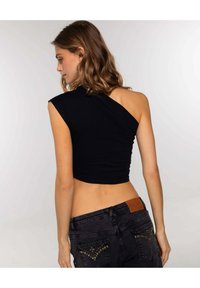 Schwarzes Crop-Top mit einseitigem Design, aus glattem Material. Getragen mit schwarzen Jeans, die mit Nietenakzenten an den Taschen versehen sind.