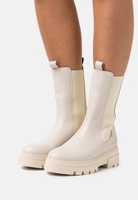Botas de tobillo de cuero beige con un acabado liso, punta rectangular, paneles laterales elásticos y suela de goma gruesa. Con acentos de lengüetas de agarre.