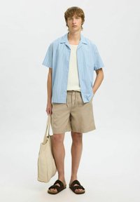 Světle modrá proužkovaná košile s krátkým rukávem, bílý top, béžové šortky a hnědé sandály. Nese béžovou tote tašku. Neformální letní outfit.
