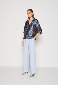 Blouse à col en V et manches courtes de couleur bleu foncé avec un motif floral métallisé, associée à un pantalon large bleu clair et des chaussures à talons argentées.