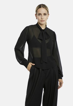 Frau mit blondem Haar, die eine transparente schwarze Bluse mit Schleife und schwarze Hose trägt, steht mit einer Hand in der Tasche vor weißem Hintergrund.