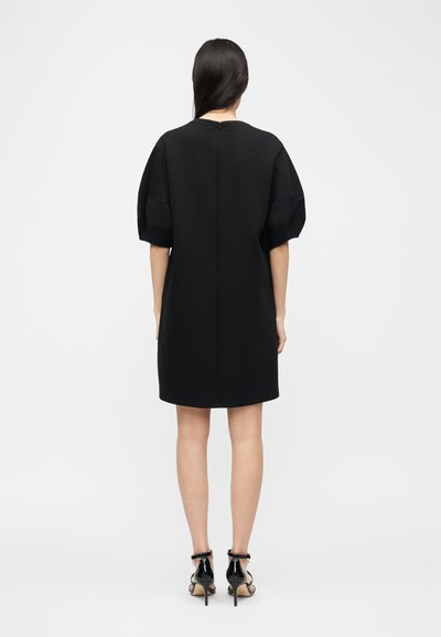 Γυναίκα με μακριά σκούρα μαλλιά φοράει μαύρο φόρεμα μήκους μέχρι τα γόνατα με κοντά μανίκια-φουσκωτά και μαύρα ψηλοτάκουνα σανδάλια, στέκεται γυρισμένη με την πλάτη.