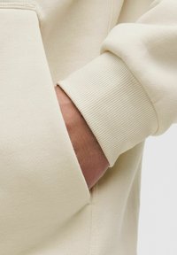 Lys beige sweatshirt med ribbet manchette. Stofbelægningen er glat, med en sidelomme og et nærbillede af ærmet og hånden.