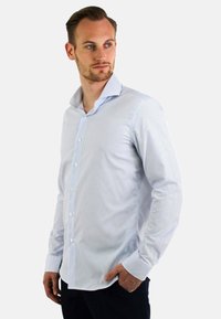 Witte blouse met blauwe verticale strepen, lange mouwen en een puntige kraag. Heeft knopen aan de voorkant en een getailleerd ontwerp.