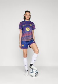 Maillot et short du FC Barcelona en bleu et violet avec des motifs ondulés. Comprend le logo Nike et un graphique Spotify. Un joueur se tient avec un ballon de football.