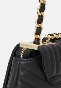 Svart vadderad läderhandväska med gul metalldetaljer. Utrustad med kedjestraps och dragkedjestängning med ett varumärkesplåt som visar "VALENTINO".