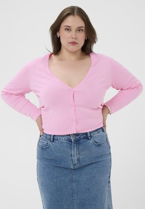 Žena so stredne dlhými hnedými vlasmi, oblečená v svetloružovom zapínacom kardigáne a modrej džínsovej sukni, stojaca s rukami v bok proti bielemu pozadiu.