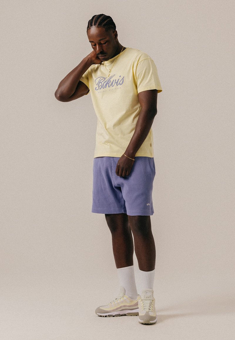 Homme aux cheveux tressés portant un t-shirt jaune à motif graphique, un short lavande, des chaussettes blanches et des baskets beiges, debout la main sur la nuque.