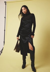 Juoda satino suknelė su šonu skilimu, uždėta ant juodos ilgos rankovės palaidinės. Papuošta juodomis aulinėmis batais iki kelių ir laikoma ruda rankine.