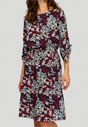 Donna che indossa un vestito al ginocchio con una fantasia floreale mista rosso, bianco, beige e nero e maniche a tre quarti legate ai polsini.