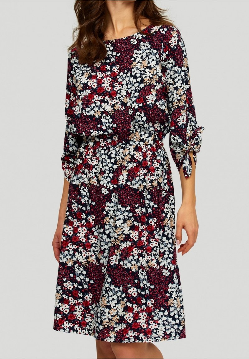 Femme portant une robe longueur genou avec un motif floral mêlé rouge, blanc, beige et noir et des manches trois-quarts nouées aux poignets.