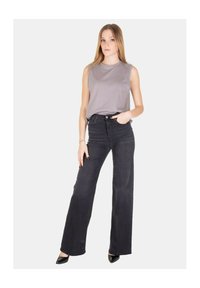 Top grigio senza maniche abbinato a jeans neri a vita alta e gamba larga. La modella è in piedi con una mano sui fianchi, indossando tacchi neri.