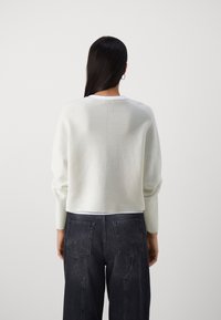 Vaaleanharmaa neulepaita, jossa on pudotetut olkapäät, pyöreä kaula-aukko ja ribatut hihansuut, yhdistettynä tummaihoisiin, korkeavyötäröisiin farkkuihin, joissa on kaksi takataskua.