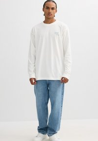 Camisa blanca de algodón de manga larga con el texto azul "Wild" en el pecho, combinada con unos vaqueros de denim azul claro sueltos y zapatillas blancas.