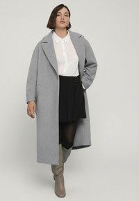 Lungo cappotto grigio in lana con colletto a punte sopra una camicia bianca, abbinato a una gonna plissettata nera e stivali grigi fino al ginocchio.