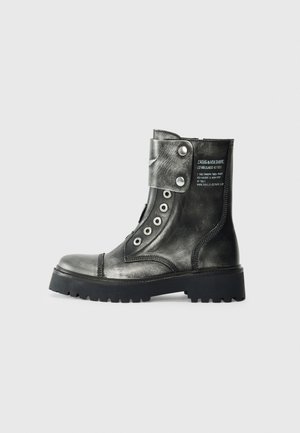 Botte de combat en cuir noir avec œillets métalliques, rabat pliable avec boutons-pression, semelle robuste et texte blanc imprimé sur le côté.