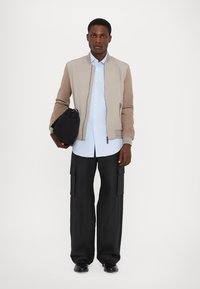 Bomberjacke in Hellgrau und Beige, knöpfbares blaues Hemd, schwarze Cargo-Hosen, hält eine schwarze Tasche, zeigt einen figurbetonten und geschichteten Look.
