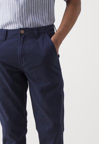 Homme portant un pantalon bleu marine avec la main dans la poche et une chemise à manches courtes rayée bleu clair et blanc sur un fond uni.