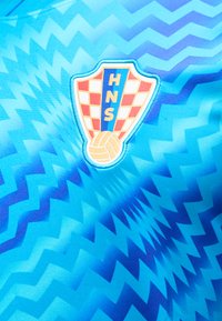 Μπλε αθλητικό ύφασμα με ζιγκ-ζαγκ μοτίβο και έμβλημα ασπίδας που δείχνει κόκκινα και λευκά καρό, τα γράμματα HNS και μια κίτρινη ποδοσφαιρική μπάλα.