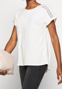 Haut blanc à manches courtes avec un col rond, orné de détails en dentelle sur les épaules et une coupe ample. Associé à une pochette rose pâle.