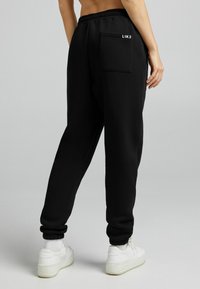 Person som bär svarta joggingbyxor med en bakficka märkt "L1K3" och vita sneakers, stående mot en enfärgad grå bakgrund.