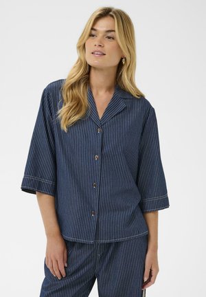 Femme aux longs cheveux blonds portant une chemise à rayures bleu marine boutonnée et un pantalon assorti, debout devant un fond clair uni.