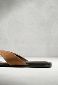 Sandale à enfiler en daim marron avec larges brides croisées et semelle plate noire sur fond gris clair.