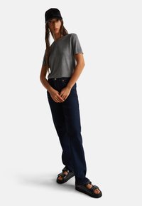 Šedé bavlněné tričko s krátkým rukávem, tmavě modré džíny rovného střihu a černé sandály s přezkou. Černá kšiltovka s logem.