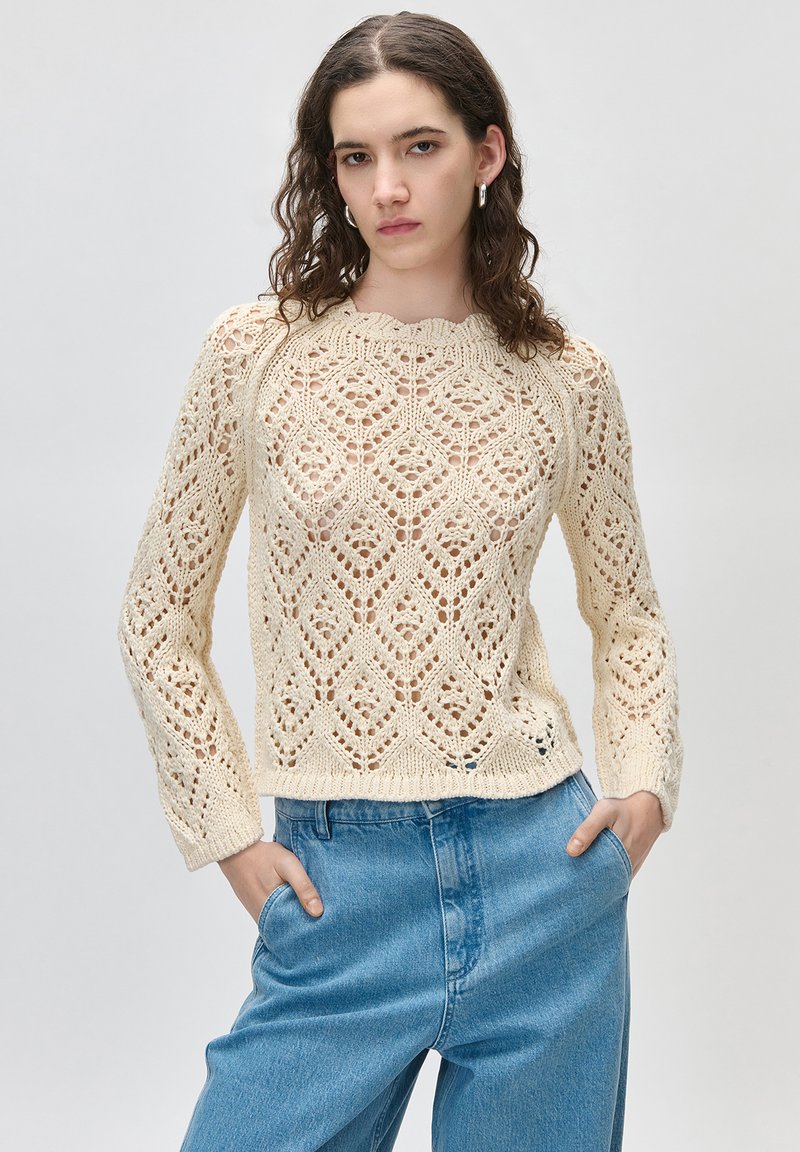 Νεαρή γυναίκα με κυματιστά καστανά μαλλιά φοράει μια εκρού πλεκτή μπλούζα και μπλε τζιν, στέκεται με τα χέρια στις τσέπες μπροστά σε απλό φόντο.