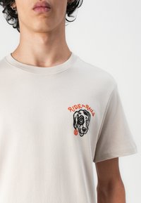 Beige kokvilnas T-krekls ar apaļu kakla izgriezumu, ar melno un oranžo grafiku, kas attēlo suņa galvu, un tekstu "RIDE the RAINS."