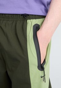 Zelené a tmavě olivové sportovní kalhoty s elastickým pasem, boční kapsou na zip a černými akcenty v oblasti kapsy.