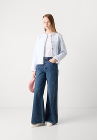 Veste matelassée bleu clair avec une texture côtelée et des boutons dorés, associée à un jean bleu foncé à jambes larges et des chaussures blanches. Tient un sac rose.