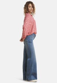 Blouse rose avec un motif de cœurs blancs, manches bouffantes ; associée à un jean évasé bleu clair et des baskets beiges. Tissu et design simples.