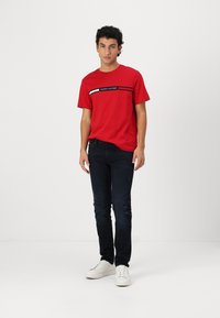 Camiseta de algodón roja con rayas horizontales en blanco y negro que presenta el texto "TOMMY HILFIGER", combinada con unos jeans oscuros y unas zapatillas blancas.