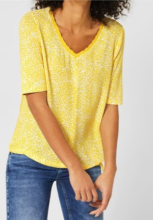 Vrouw draagt een geel patroon V-hals shirt met korte mouwen en blauwe spijkerbroek met handen geklemd voor zich tegen een witte achtergrond.