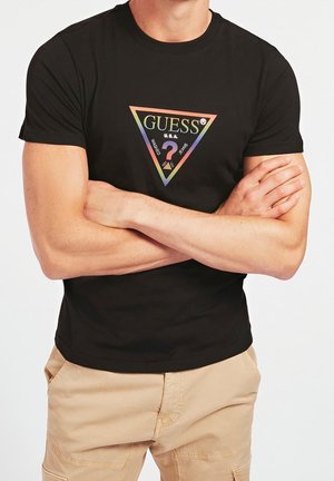 Man met over elkaar geslagen armen, draagt een zwart Guess T-shirt met regenboog driehoek logo en beige broek tegen een witte achtergrond.