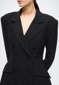 Černý dvouřadý blejzr s hlubokým výstřihem do V, strukturovanými rameny, velkými knoflíky a dvěma předními kapsami s klopami. Hladká textura látky.
