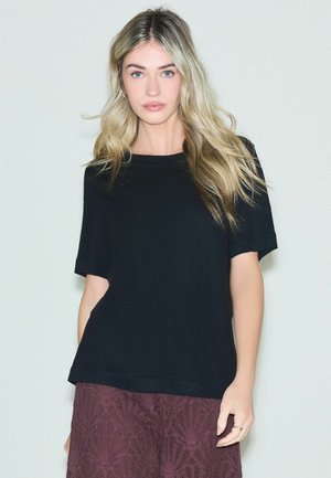 Young woman with long blonde hair wearing a black short-sleeve top and textured dark purple skirt, standing against a plain light background.