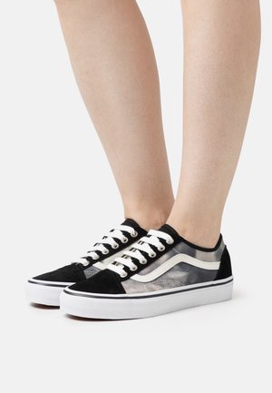 Des jambes portant des baskets à lacets noires et grises avec des semelles blanches et une bande blanche sur le côté, debout sur un fond clair uni.