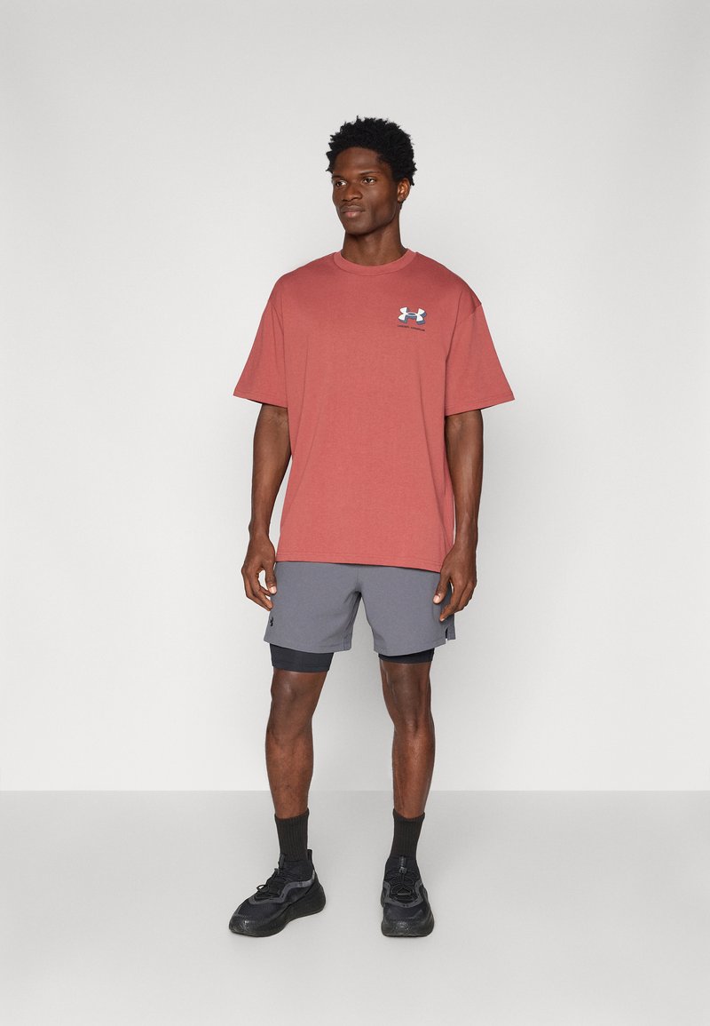 Camiseta de manga corta color óxido con un logo azul, combinada con pantalones cortos grises. El portador está de pie, vistiendo zapatillas deportivas negras y calcetines.