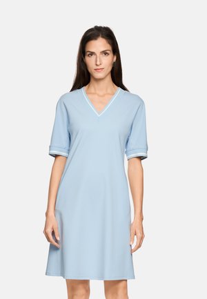 Frau trägt ein hellblaues, kurzärmeliges Kleid mit V-Ausschnitt und weißen Streifen am Kragen und an den Ärmelbündchen, steht vor einem einfarbigen Hintergrund.