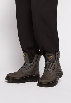 Person trägt schwarze Hose und graue hohe Dr. Martens Stiefel mit dicken schwarzen Sohlen und gemusterten Schnürsenkeln, steht auf einer weißen Oberfläche.
